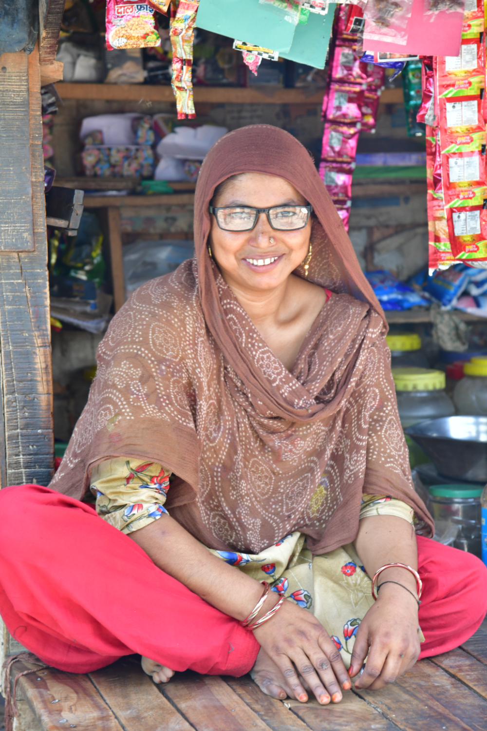 A photo of a woman sitting in front of a shop, smiling at the camera. She is wearing glasses that she acquired in a Peek-powered eye health programme in India.