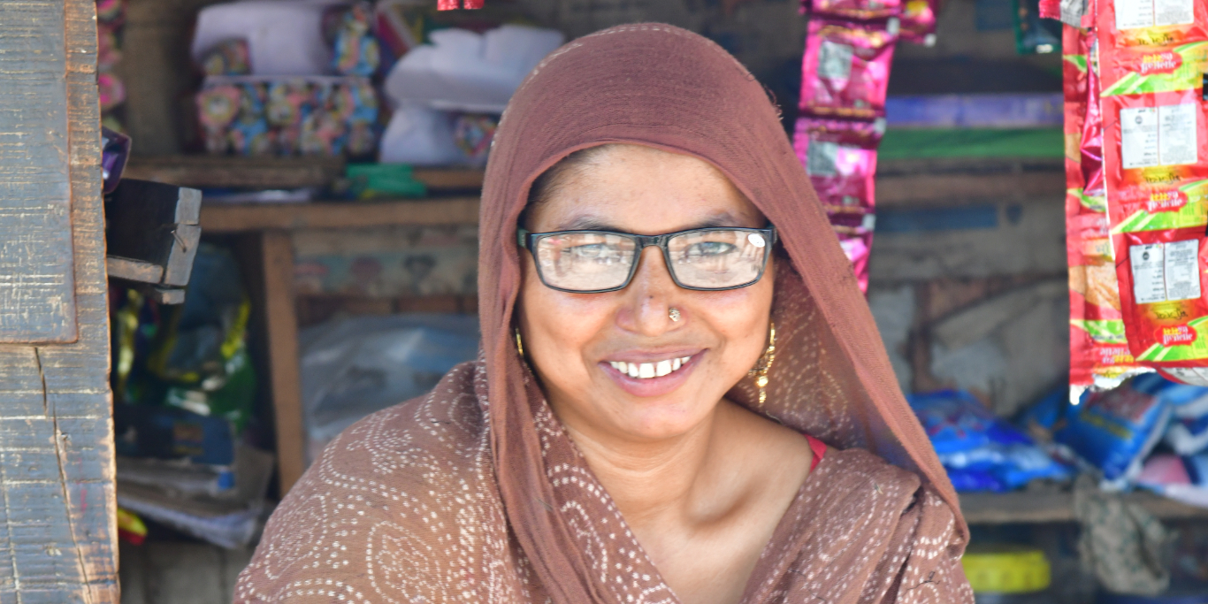 A photo of a woman sitting in front of a shop, smiling at the camera. She is wearing glasses that she acquired in a Peek-powered eye health programme in India.