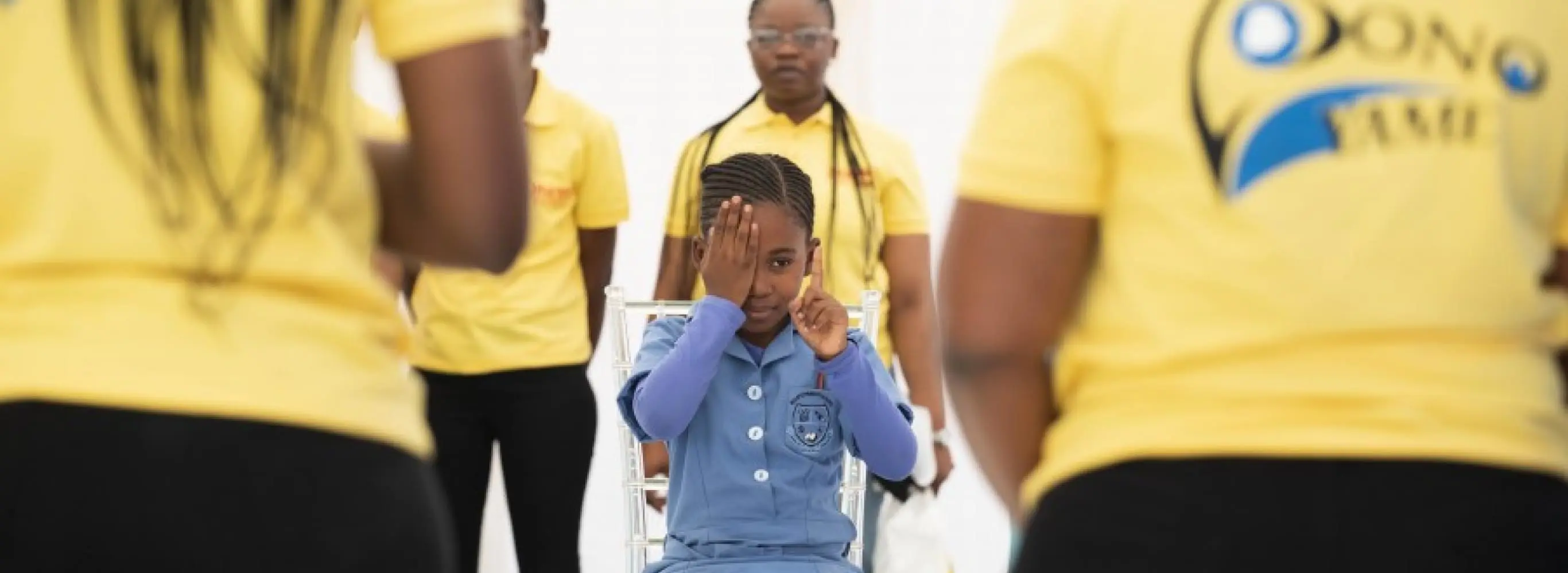 A school girl covers one of her eyes with her hands as she has her vision screened in a primary school in Botswana. The screeners, wearing yellow Pono Yame branded tshirts, face away from the camera.