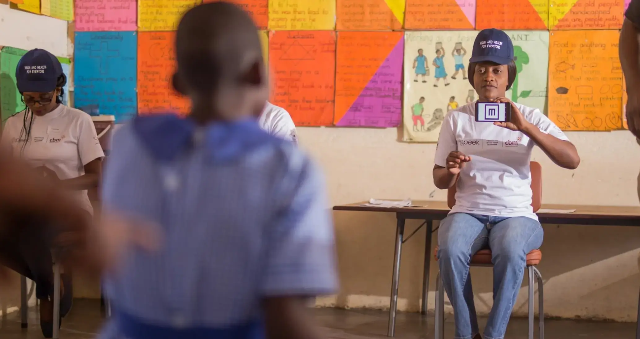 A woman uses a smartphone with Peek technology (an E symbol from a vision test app) to test the vision of a child at a school in Zimbabwe. She wears a hat that says 'One School at a Time' and is sitting in front of a colourful classroom background.