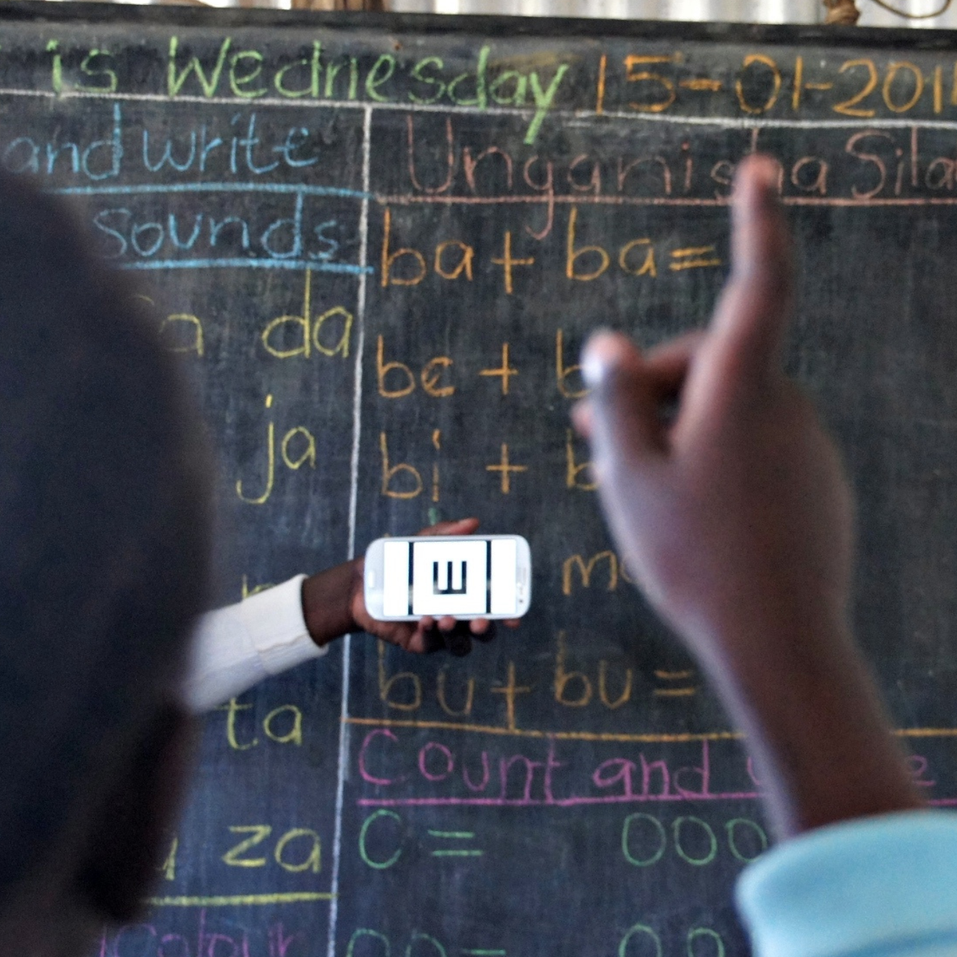 A woman holds the Peek Vision screening app in front of a blackboard in the background. In the foreground a child's hand is raised as they indicate which way the tumbling 'E' on the screen is facing as part of their vision test.