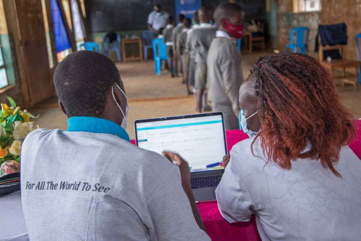 Two people look at a laptop displaying Peek Vision software for programme monitoring. They are sitting in a classroom in a school in Kenya.