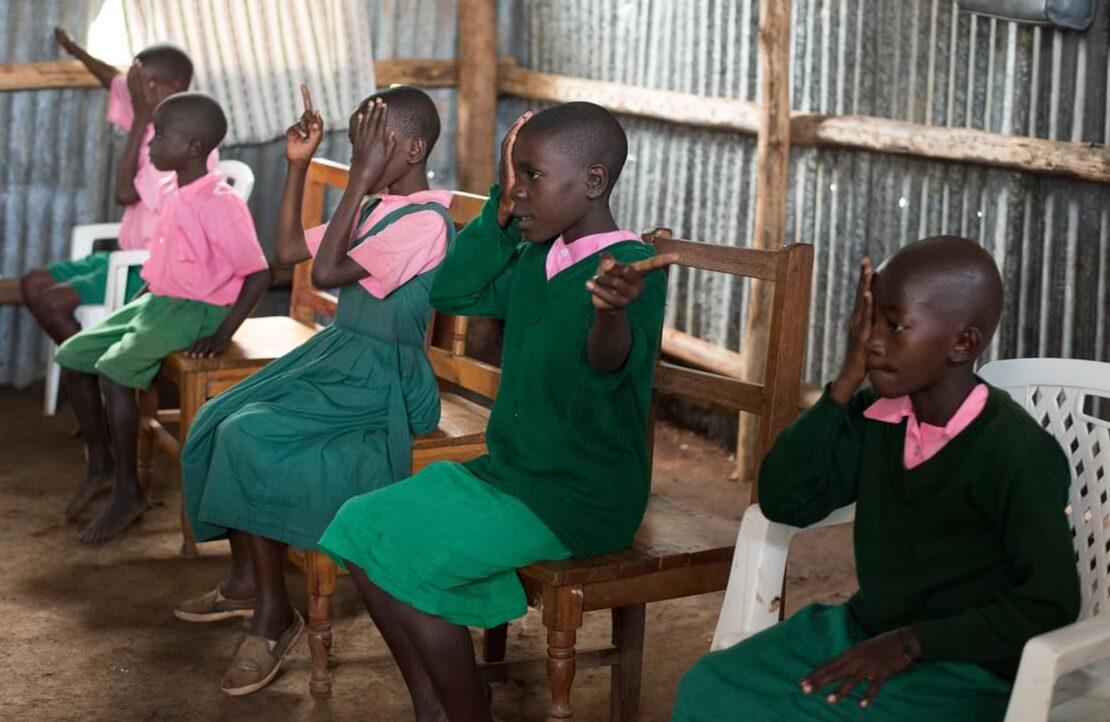 School children in Kenya sit in a row, using their hands to cover one eye as part of a vision screening session.