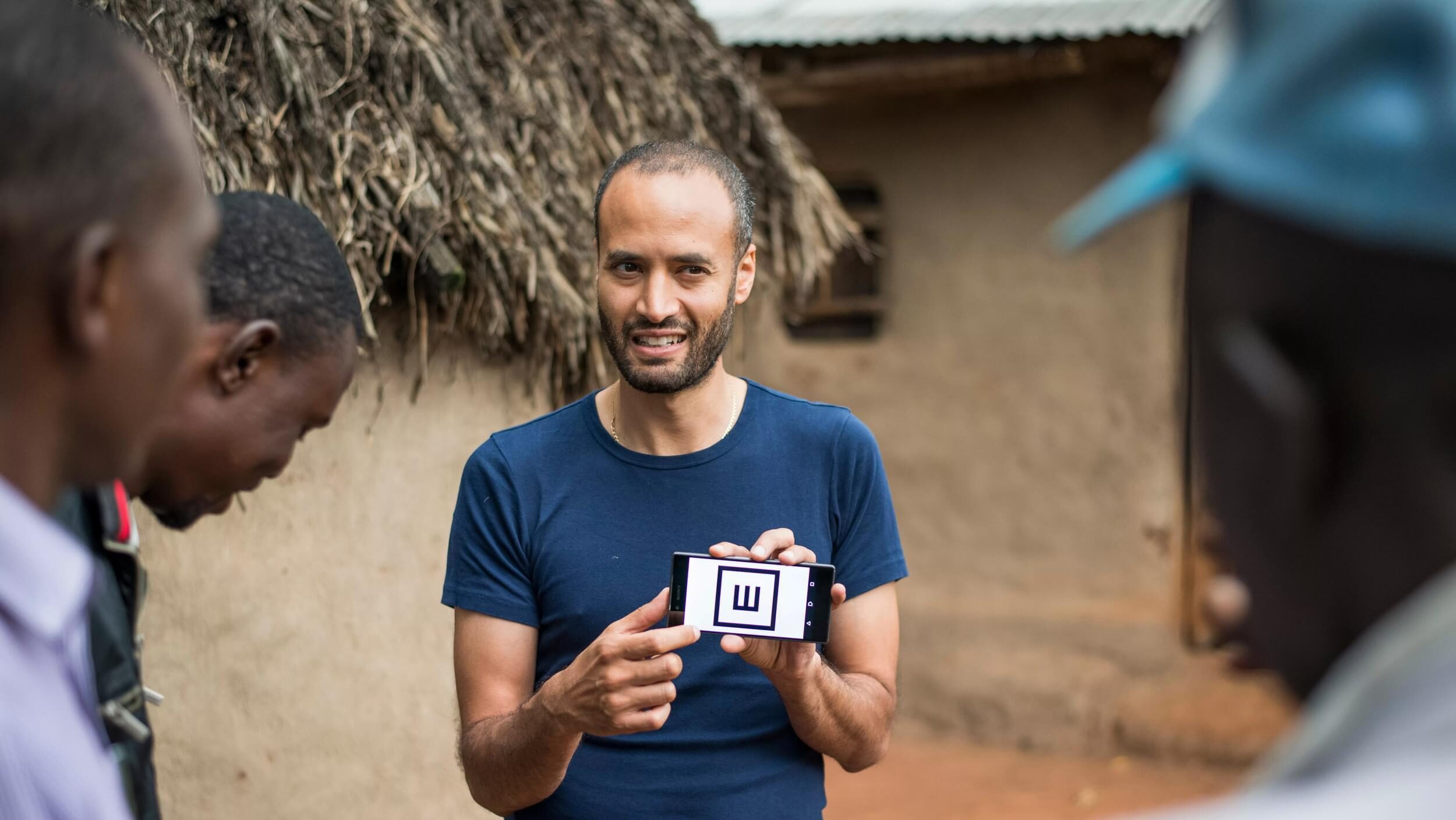 Peek CEO, Andrew Bastawrous, holds up a phone displaying the Peek app vision test with 'tumbling E'. He shows this to a group of men in rural Kenya.