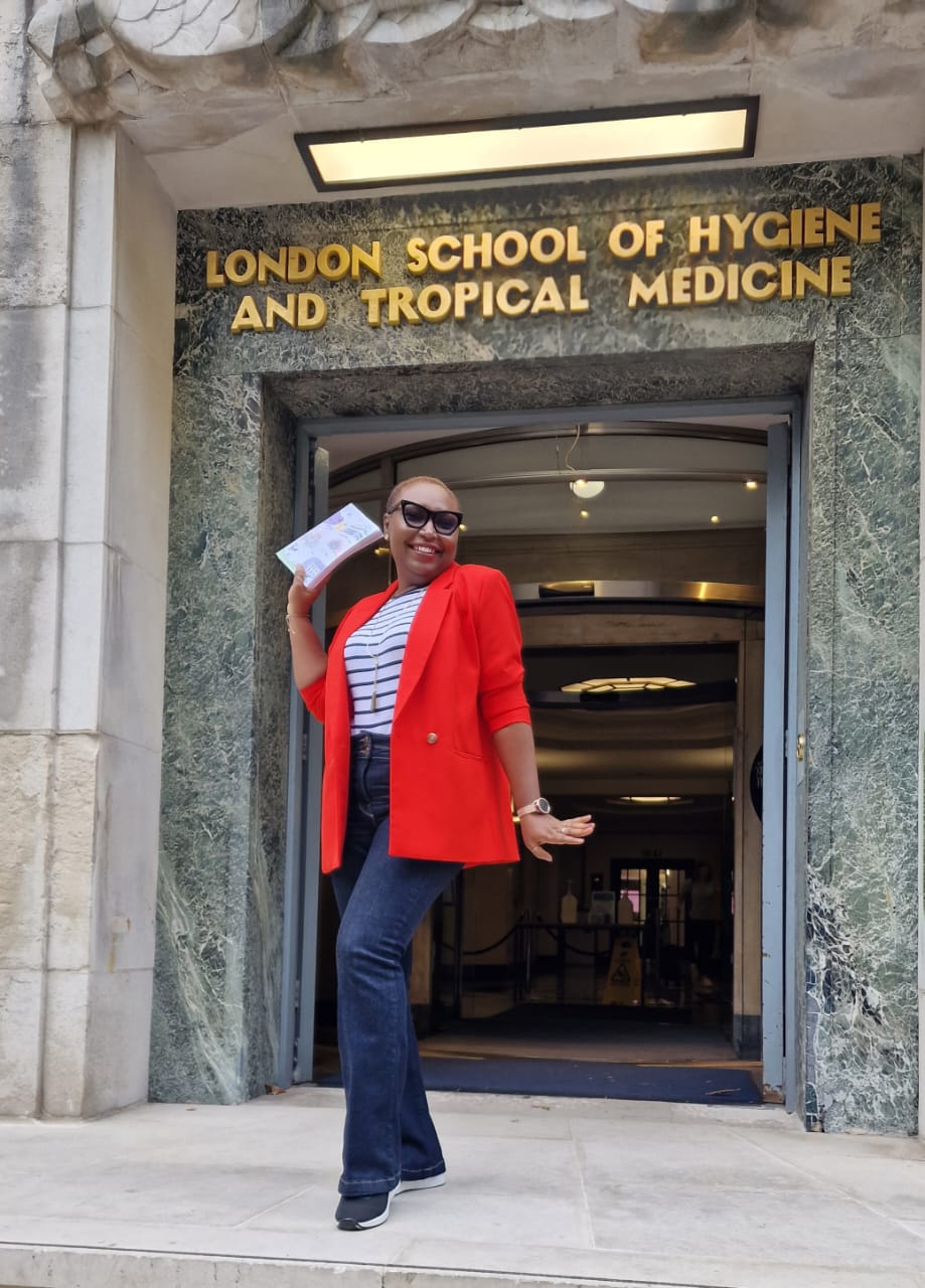 Dr Oteri Okolo stands in front of the London School of Hygiene and Tropical Medicine. She is smiling and is holding a certificate of graduation in her hand.