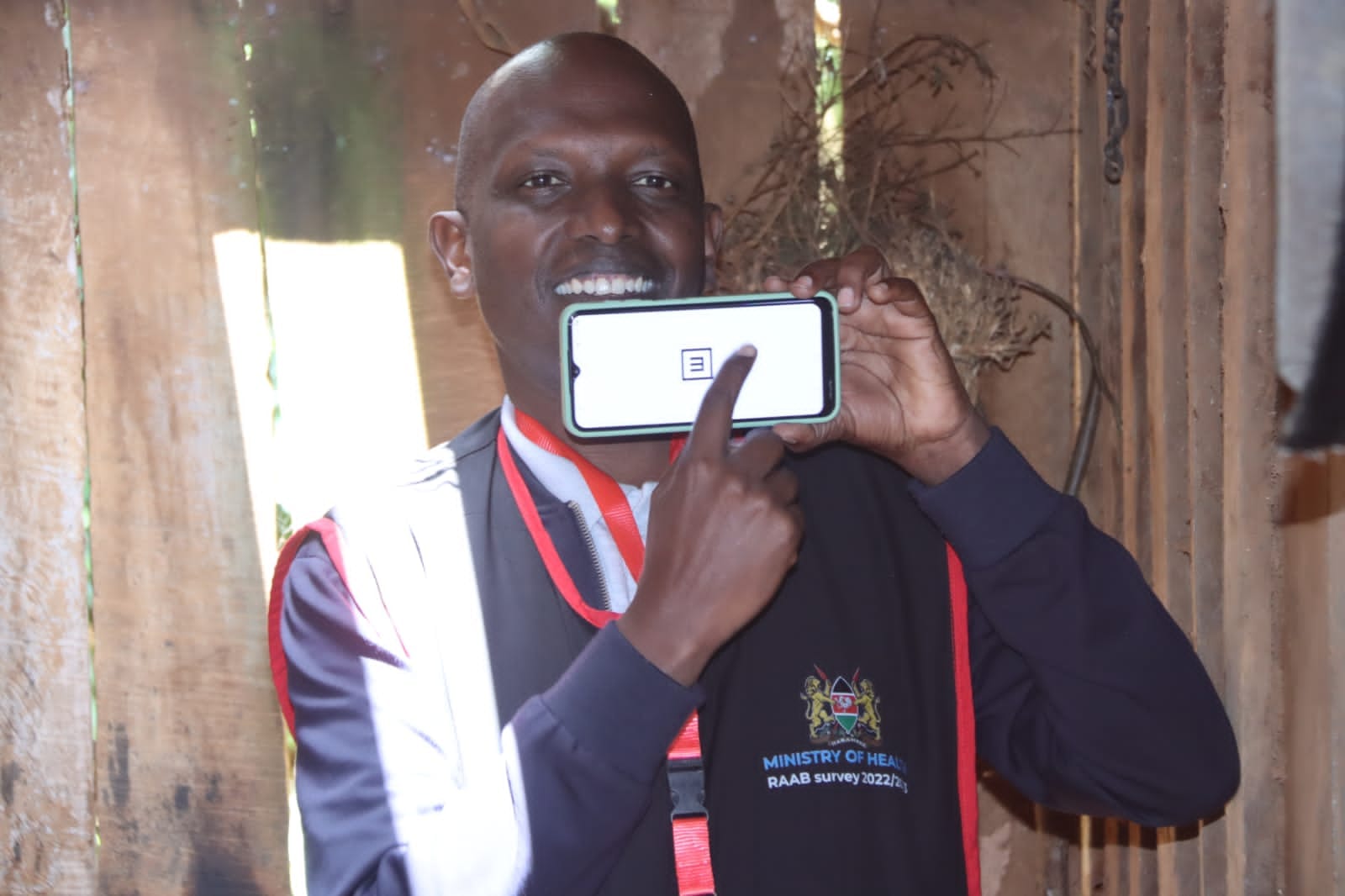 Dr Hillary Rono from Peek holds up a phone displaying the Peek Vision visual acuity test. He is swiping the phone and he wears a vest with the Ministry of Health Kenya logo which also says RAAB survey 2022/2023