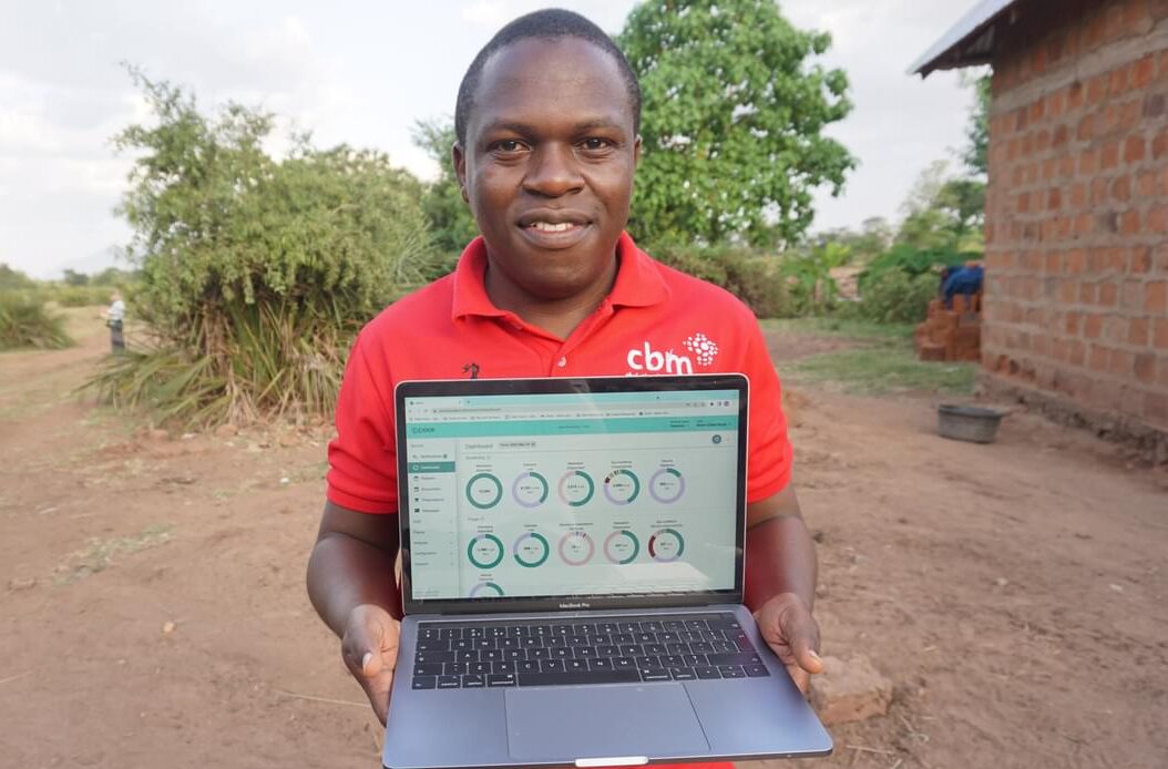 A man wearing a CBM tshirt stands holding a laptop displaying the Peek Admin dashboard.