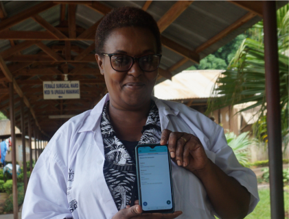 Dr Naomi Damas Shayo wearing a doctors' coat and standing in the outdoor leafy grounds of the Mawenzi Hospital in Kilimanjaro region of Tanzania. She holds the Peek Vision app opened to a data capture function.