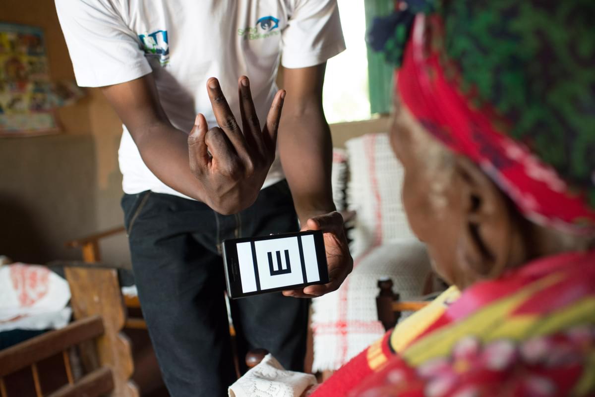 A man holds a phone with the Peek vision test app displaying the 'tumbling E'. A woman faces the phone.