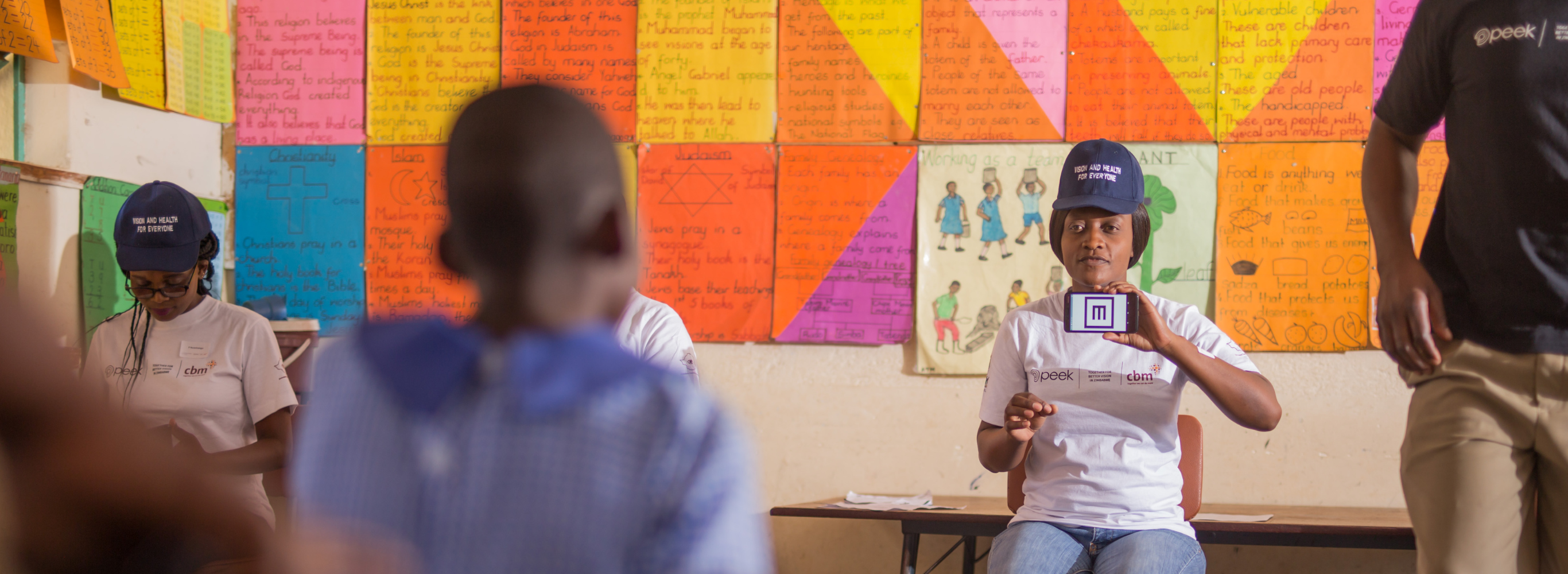 A woman uses a smartphone with Peek technology (an E symbol from a vision test app) to test the vision of a child at a school in Zimbabwe. She wears a hat that says 'One School at a Time' and is sitting in front of a colourful background.