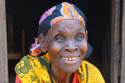 An elderly woman, Teodosia, smiles on the porch of her home in an rual village in Tanzania. She is wearing traditional African dress.
