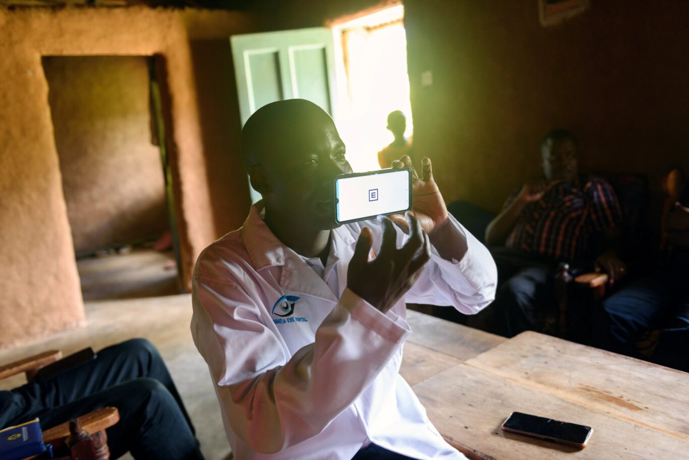 A community health assistant sits inside a home in Kenya conducting a vision screening. He holds up a phone displaying the Peek visual acuity test.