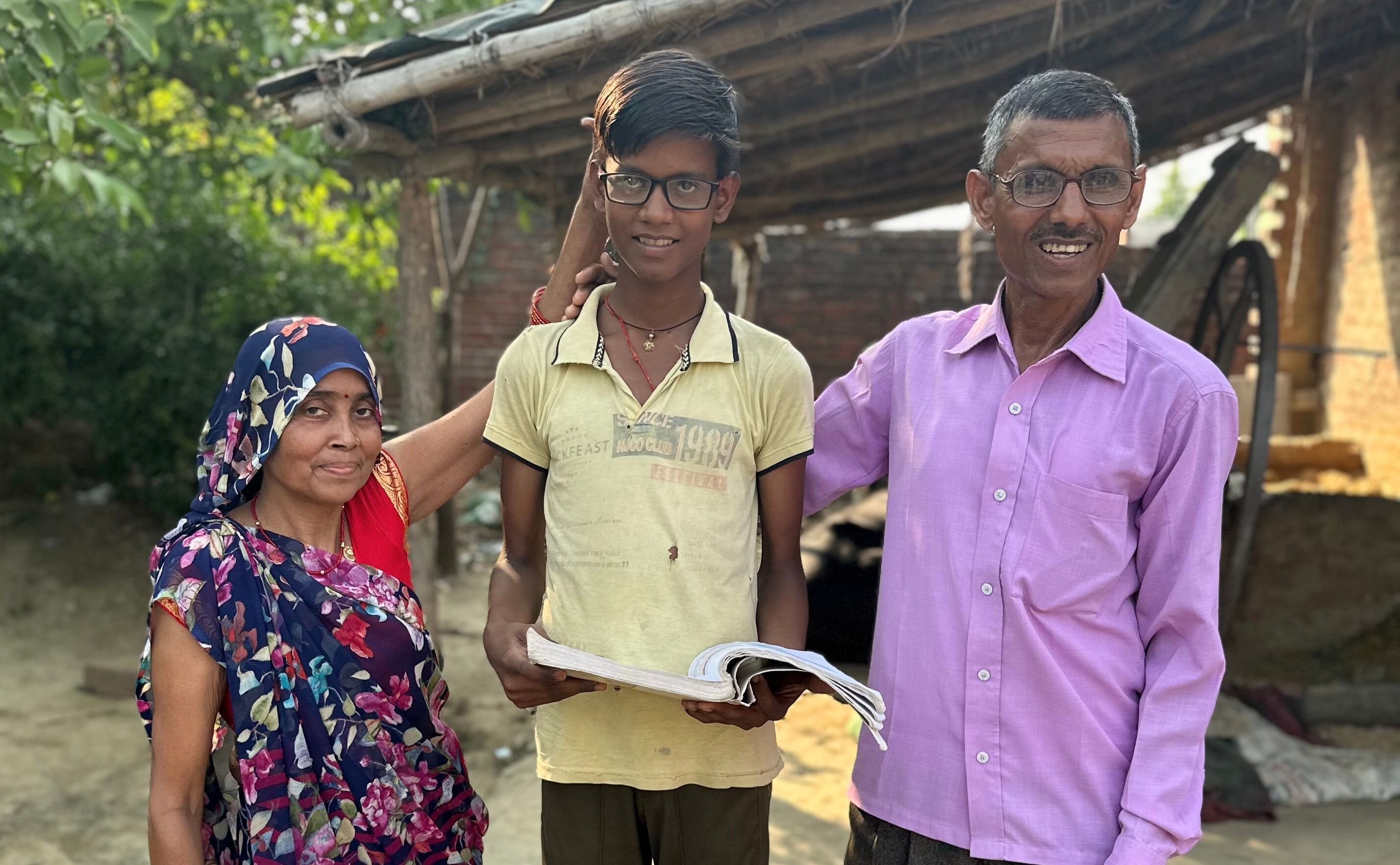 14 year old Ayush stands smiling, wearing his glasses and holding a school text book. His parents stand either side of him smiling and his mother touches his head.