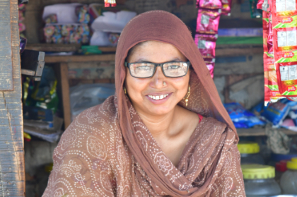 A photo of a woman sitting in front of a shop, smiling at the camera. She is wearing glasses that she acquired in a Peek-powered eye health programme in India.