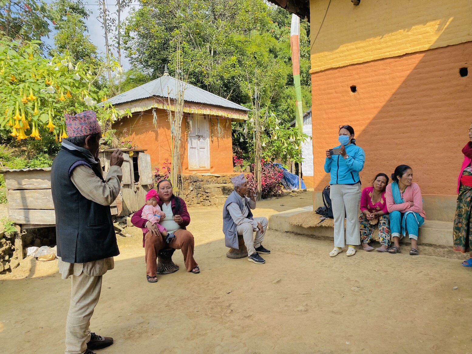 A man in a village in Nepal stands outside facing a woman conducting vision screening using a smartphone, while other community members look on.