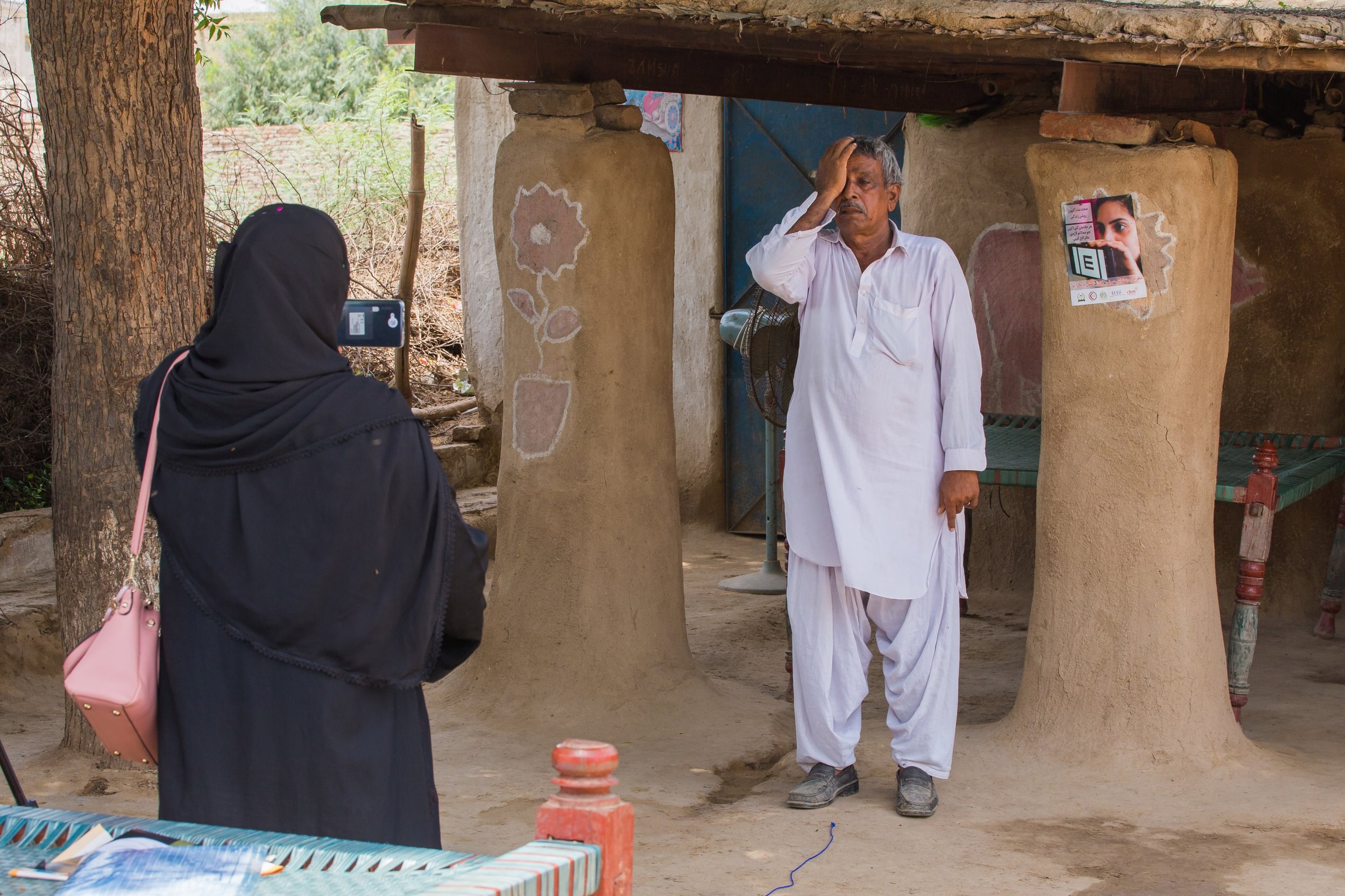 A man in rural Pakistan stands near his home and covers one eye as a Lady Health Worker conducts a vision test using the Peek app on a phone.